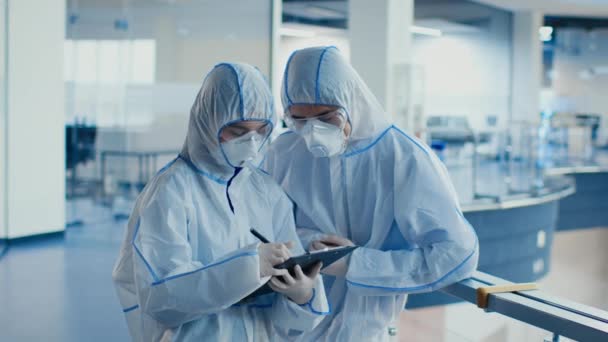Dos trabajadores médicos tomando notas con trajes protectores de materiales peligrosos en interiores — Vídeo de stock