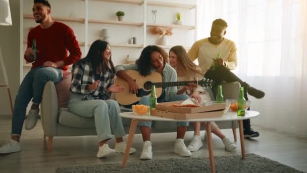 Lindos jóvenes amigos diversos tocando la guitarra y cantando canciones juntos, teniendo fiesta en casa, sentado en el sofá en la sala de estar — Vídeos de Stock