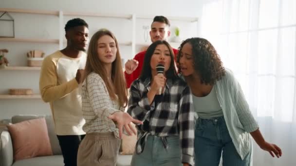 Fiesta de karaoke en casa. Grupo de mujeres multiétnicas felices cantando juntas en el micrófono, chicos bailando en el fondo — Vídeo de stock