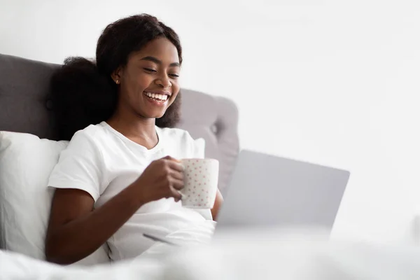 Νεαρή γυναίκα πληκτρολογώντας στο laptop και απολαμβάνοντας καφέ στο κρεβάτι — Φωτογραφία Αρχείου