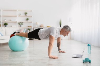 Evde eğitim. Atletik olgun bir adam dizüstü bilgisayarının önünde spor yapıyor, evden online fitness dersi alıyor.