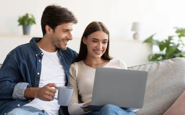 Посміхаючись, привабливий молодий чоловік з чашкою дивиться на дружину з ноутбуком у вітальні. — стокове фото
