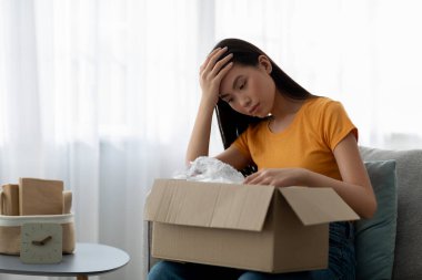 Yanlış paket. Paketleri açan Asyalı kadın oturma odasındaki koltukta oturan kötü teslimat servisinden şok olmuş.