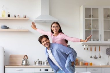 Gülümseyen yakışıklı Avrupalı koca, sıradan bir şekilde karısını sırtında taşıyor ve İskandinav mutfağında eğleniyor.