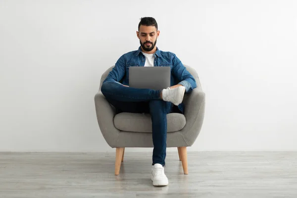 Millennial uomo arabo seduto in poltrona, utilizzando il computer portatile per il lavoro online o la comunicazione contro parete studio bianco — Foto Stock