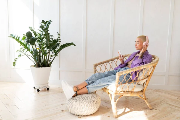 Χαλαρή γυναίκα που φοράει ασύρματα ακουστικά και χρησιμοποιεί smartphone, κάθεται σε ψάθινη καρέκλα στο σπίτι, ελεύθερος χώρος — Φωτογραφία Αρχείου
