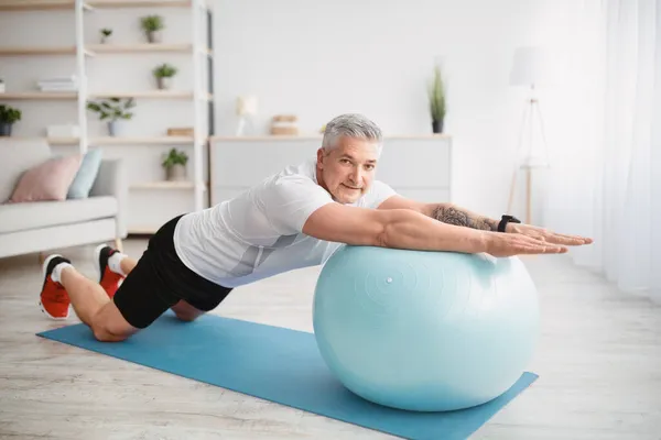 Concepto de entrenamiento en casa. Hombre mayor atlético haciendo ejercicio con pelota de fitness, ejercitando los músculos del núcleo en casa — Foto de Stock