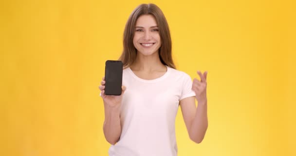 Grande aplicação móvel. Jovem mulher alegre mostrando celular com tela em branco e apontando para ele com o dedo — Vídeo de Stock