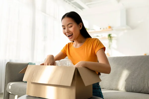 Online vásárlás, öröm a vásárlás. Boldog fiatal ázsiai hölgy kinyitja a kartondobozt és belenéz — Stock Fotó
