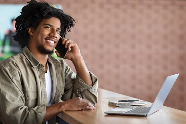 Negro freelancer chico hablando en el teléfono celular y trabajando con el ordenador portátil en café —  Fotos de Stock