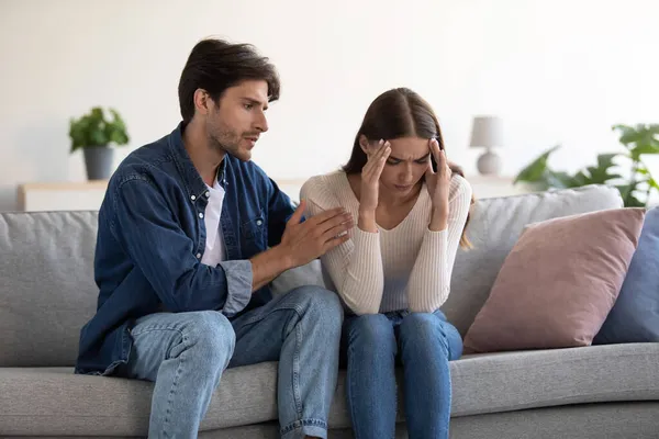 Young caucasian couple sit on sofa, husband calms upset wife at home interior