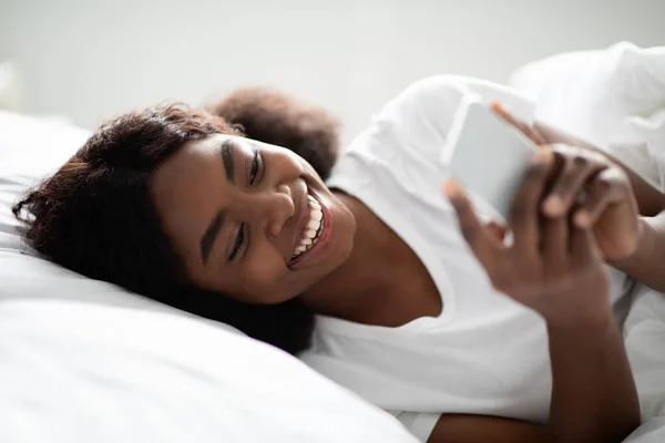 Alegre mujer afroamericana utilizando el teléfono celular en la cama —  Fotos de Stock