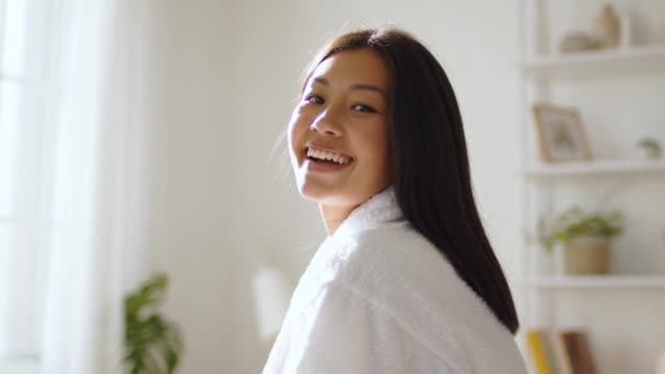 Fiatal békés koreai nő fehér fürdőköpenyben mosolyog a kamerába, pózol otthon reggel, napfény, lassított felvétel — Stock videók