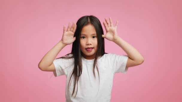 Азиатская девочка Гримасинг держа руки возле храмов, розовый фон — стоковое видео