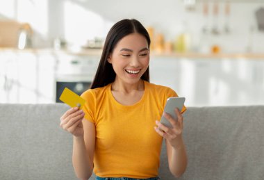 Çevrimiçi ödeme kavramı. Kredi kartı ve akıllı telefon tutan heyecanlı Asyalı kadın, koltukta oturuyor, internetten alışveriş yapıyor.