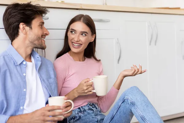 Boldog Y-generációs fehér pár csészékkel, kávézgatnak reggel, leülnek a földre, szabadidőben beszélgetnek. — Stock Fotó