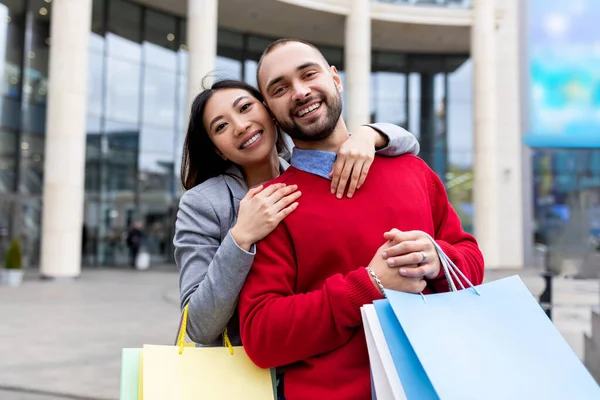 Aanhankelijk interraciaal paar met shopper tassen glimlachen en knuffelen in de buurt van groot winkelcentrum, buiten — Stockfoto