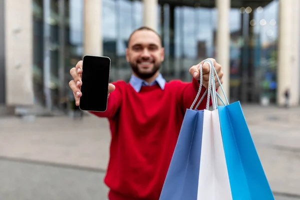 Junger Kerl mit hellen Geschenktüten zeigt Smartphone mit leerem Bildschirm, Online-Shopping in der Nähe der City Mall, Attrappe — Stockfoto