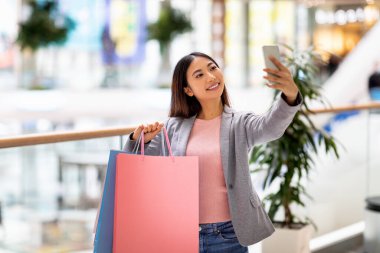 Alışveriş poşetleriyle güzel bir Asyalı kadın alışveriş merkezinde akıllı telefondan selfie çekiyor, marka logosu için maket yapıyor.