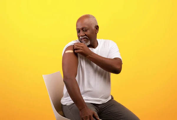 Happy elderly black man showing band aid on shoulder after coronavirus vaccination over orange studio background
