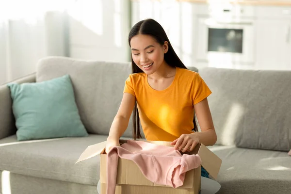 Szállítószolgálat. Pozitív ázsiai hölgy kipakol csomag doboz vásárlás, ül a kanapén, elégedett a megrendelt áruk — Stock Fotó