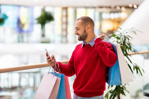 Gelukkige jongeman met kleurrijke geschenkzakjes met behulp van een mobiele telefoon in het winkelcentrum, het controleren van de boodschappenlijst, het hebben van video bellen, kopiëren ruimte — Stockfoto