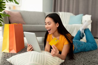 Aşırı neşeli Asyalı kadın tablet bilgisayar ve kredi kartıyla yerde yatıyor, internetten alışveriş yapıyor ve satışa duygusal tepki veriyor.