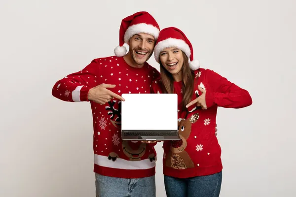 Cheerful Young Couple Wearing Santa Hats Pointing At Laptop With Blank Screen — Stock Photo, Image