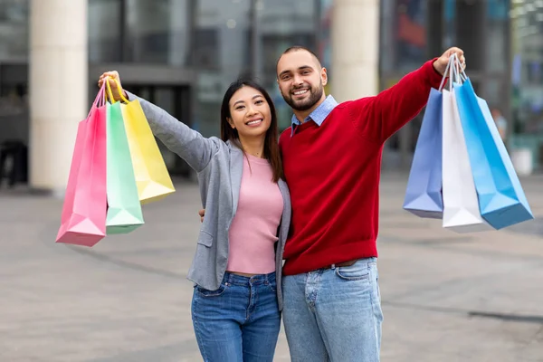 Tysiącletnia, zróżnicowana para stojąca obok centrum handlowego, trzymająca torby na zakupy w wyciągniętych dłoniach, uśmiechająca się do kamery — Zdjęcie stockowe