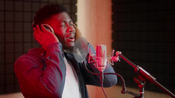 Producción musical. negro chico usando auriculares cantando canción a micrófono en estudio — Vídeos de Stock