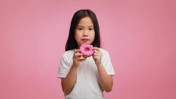 Корейська дівчинка їсть пончик посміхаючись позує над рожевим тлом — стокове відео