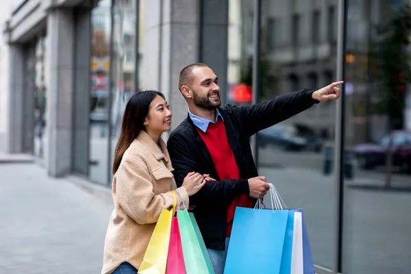 Pozytywna para międzyrasowa spacerująca ulicą z papierowymi torbami, wskazująca na nowe okno sklepu, robiąca razem zakupy — Zdjęcie stockowe
