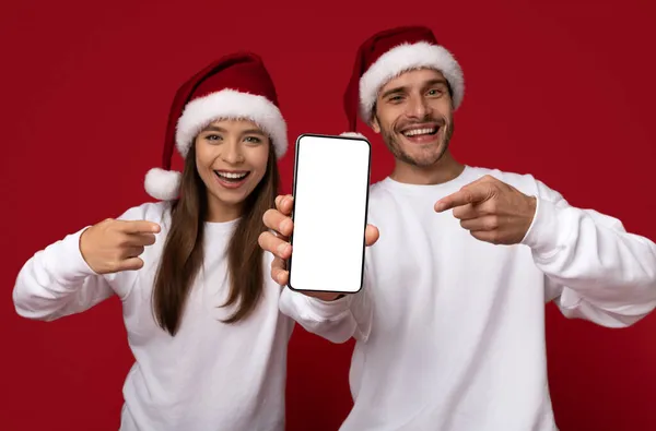 Xmas Ad. Happy Young Couple In Santa Hats Pointing At Blank Smartphone — Stock Photo, Image