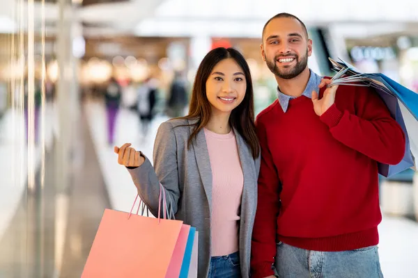 Szczęśliwa młoda wielorasowa para z kolorowymi torbami shopper stojącymi razem po zakupach w centrum handlowym, przestrzeń kopiująca — Zdjęcie stockowe