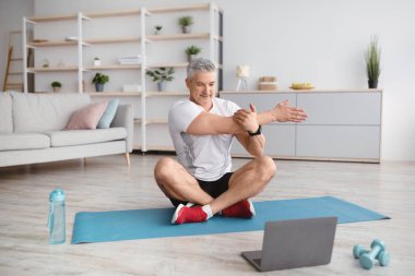 Atletik olgun bir adam evde çalışırken laptopun önünde yoga yapıyor, pilates yapıyor, paspasın üzerinde geriniyor.