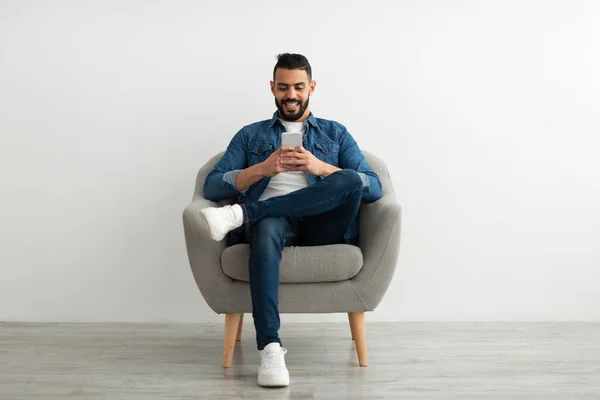 Glada unga arabiska man som använder smartphone, har online chat, sitter i fåtölj, bläddrar sociala medier — Stockfoto
