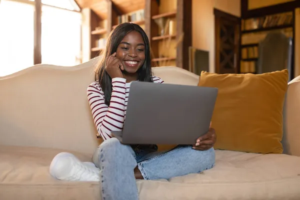 Online-Job, freiberufliche Tätigkeit, Fernausbildung. Junge schwarze Frau sitzt mit Laptop auf Sofa, arbeitet oder lernt von zu Hause — Stockfoto