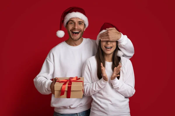 산타 모자를 쓴 흥분 한 청년 이 여자 친구의 눈을 가리고 선물을 주다 — 스톡 사진