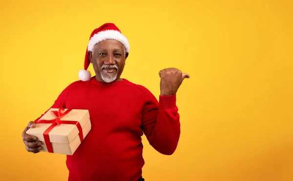 Alegre anciano negro sosteniendo regalo de Navidad, señalando a un lado el espacio en blanco en fondo de estudio naranja — Foto de Stock