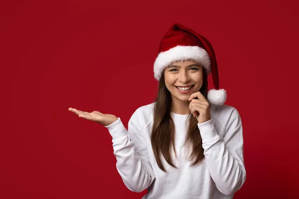 Vánoční nabídka. Veselá mladá žena v Santa Klobouk držení neviditelný objekt na otevřené dlani — Stock fotografie