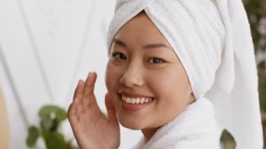 Sağlık konsepti. Kafasında havlu olan neşeli Asyalı kadın kameraya gülümsüyor, yüzüne nemlendirici krem sürüyor.