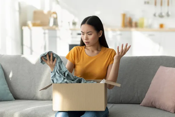 Unzufriedene junge asiatische Frau öffnet Schachtel und betrachtet falsch gelieferte Ware, unglücklich mit erhaltenem Hemd — Stockfoto