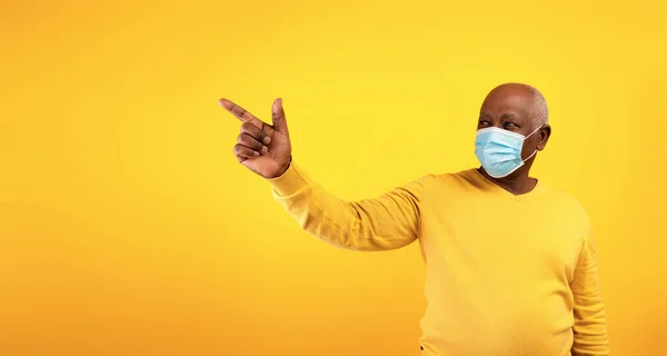 Äldre svart man i kirurgisk ansiktsmask pekar på tomt utrymme för text eller annons över orange studio bakgrund, banner — Stockfoto