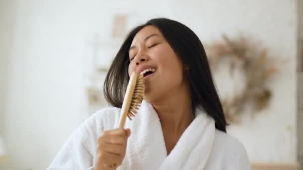 Cura della bellezza e divertimento. Giovane donna asiatica positiva che indossa accappatoio cantare con spazzola per capelli, scherzare in giro a casa — Video Stock