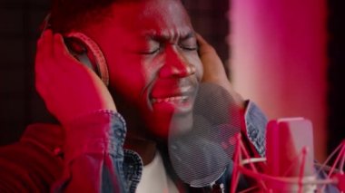 Müzik stüdyosunda profesyonel mikrofona şarkı söyleyen kulaklık takan duygusal siyahi adam.