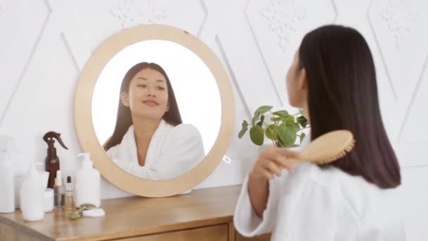 Rutina de belleza cotidiana. joven atractivo pacífico asiático señora peinando su cabello con cepillo, mirando espejo en dormitorio — Vídeo de stock