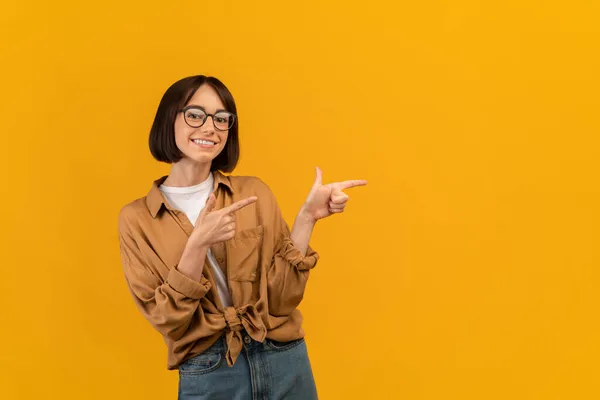 Портрет щасливої молодої леді, що вказує на два пальці позаду під час копіювання простору реклами вашого тексту, жовтого фону — стокове фото