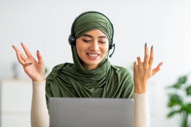 Başörtülü, kulaklık takan, evde dizüstü bilgisayar başında buluşan genç, pozitif Arap bir kadın.