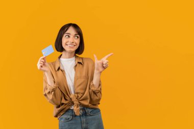 Kredi kartıyla heyecanlı beyaz kadın sarı stüdyo arka planında boş bir yere bakıyor.