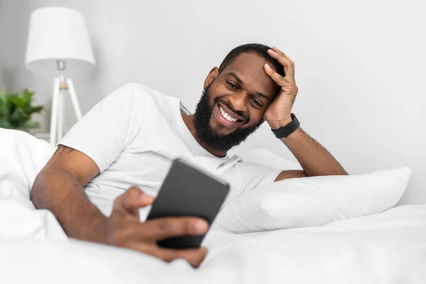 Χαρούμενος νεαρός Αφροαμερικάνος με γενειάδα κοιτάζει το smartphone, ξαπλώνει στο λευκό κρεβάτι. — Φωτογραφία Αρχείου